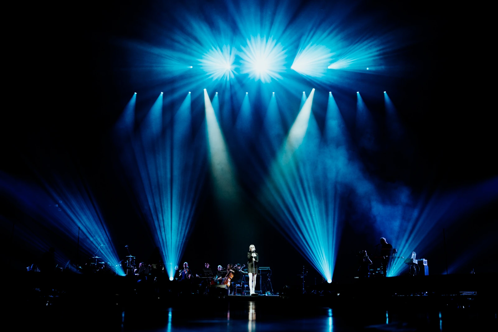 A singer and orchestra beneath sweeping blue beams of light
